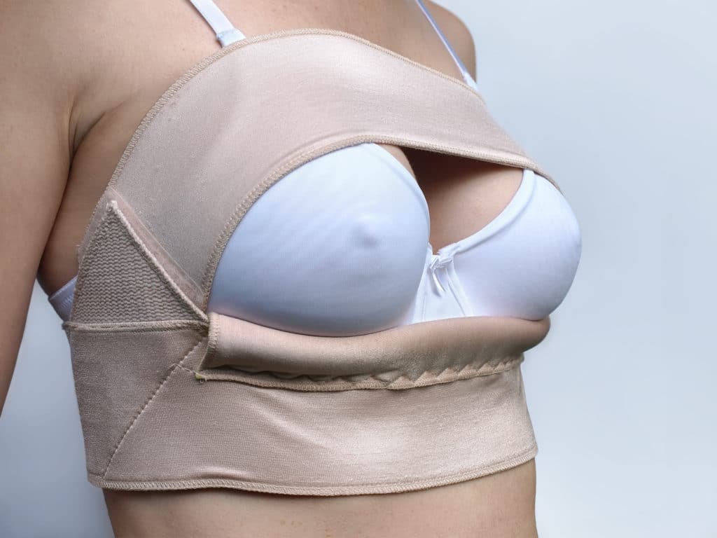 An image of a woman wearing a white bra with a breast garment: double strap bandeau, that provides post-surgical support following breast reconstruction surgery.